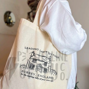 May include: Cream-colored tote bag with a black illustration of a cottage and the text "LEADING LADY COTTAGE SURREY, ENGLAND be the leading lady of your own life". The bag is carried over the shoulder.