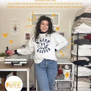 May include: A woman wearing a white sweatshirt with the text "MAJOR WEEPER" in blue lettering. She is standing in front of a white table with a heat press and a shelf unit with stacks of white t-shirts. The text "POWERLINE SUPPLY" is visible in the lower left corner of the image.