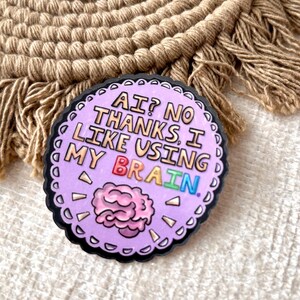 May include: A round, lavender-colored pin with a black border. The pin features the text "AI? NO THANKS, I LIKE USING MY BRAIN" in white, with the word "BRAIN" in rainbow colors. A pink brain graphic is also included.