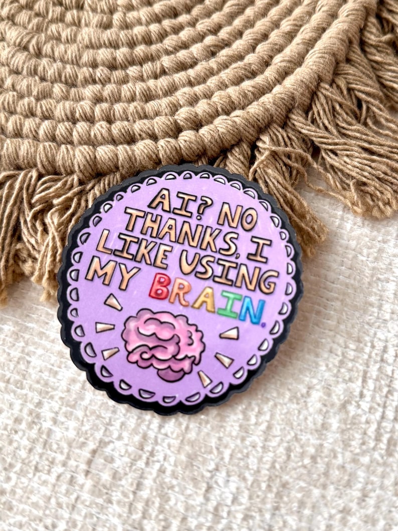 May include: A round, lavender-colored pin with a black border. The pin features the text "AI? NO THANKS, I LIKE USING MY BRAIN" in white, with the word "BRAIN" in rainbow colors. A pink brain graphic is also included.