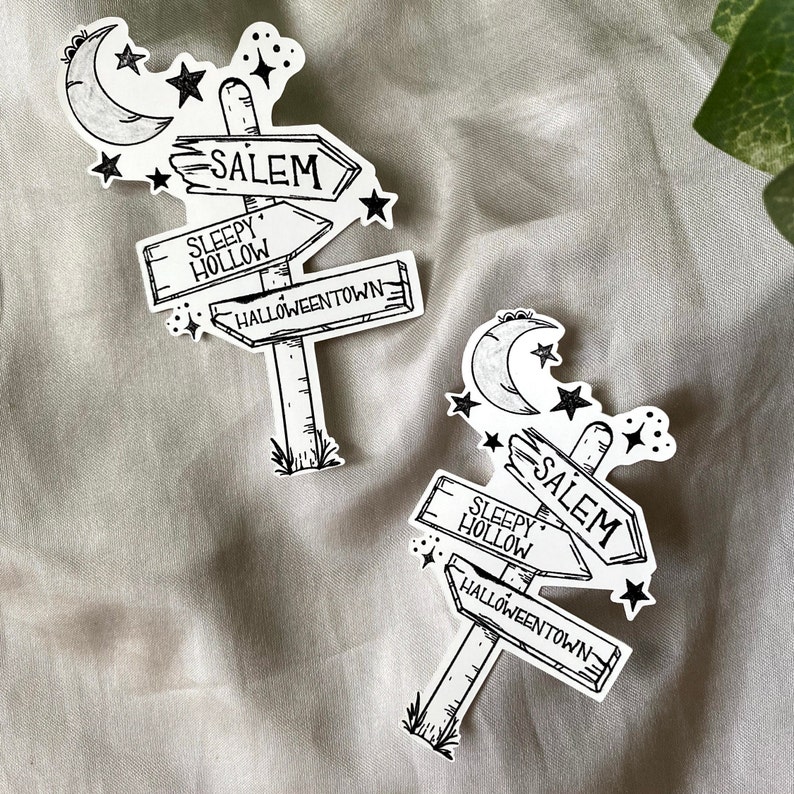 May include: Two black and white illustrations of a signpost with three arrows pointing to different locations. The signpost has a crescent moon and stars above it. The arrows point to Salem, Sleepy Hollow, and Halloweentown.