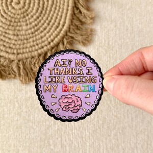 May include: A round sticker with a black scalloped border and the text "AI? NO THANKS, I LIKE USING MY BRAIN." The sticker has a light purple background and a pink brain graphic. The sticker is held by a hand.