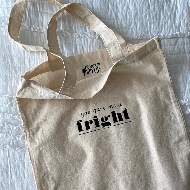 Op de afbeelding: Een beige canvas draagtas met zwarte handvatten en de tekst "you gave me a fright" gedrukt op de voorkant. De tas heeft ook een klein zwart label met de tekst "Powerline Supply."