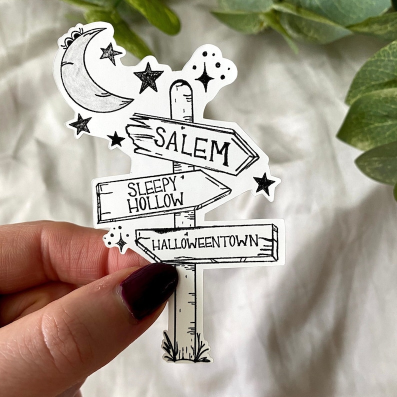 May include: Black and white illustration of a signpost with three directional arrows. The arrows point to Salem, Sleepy Hollow, and Halloweentown. The signpost is decorated with a crescent moon and stars.