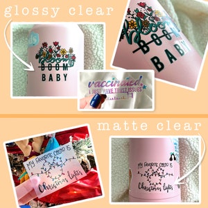 May include: Two images showing the difference between glossy and matte clear vinyl stickers. The glossy sticker is on a pink water bottle and says "Bloom Boom Baby" with flowers. The matte sticker is on a pink water bottle and says "My favorite color is Christmas lights" with a string of lights.