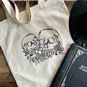 May include: A white canvas tote bag with black line art of two skulls with a banner that reads "Till Death". The skulls are surrounded by flowers.
