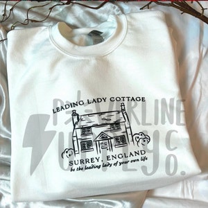 May include: White sweatshirt with a black graphic of a cottage with the text "LEADING LADY COTTAGE SURREY, ENGLAND be the leading lady of your own life"
