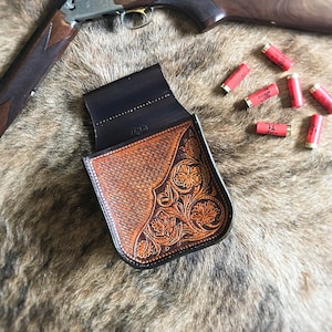 Puede incluir: Un portacartuchos de escopeta de cuero marrón con un diseño floral tallado. El portacartuchos está sobre una superficie de piel sintética marrón con una escopeta y cartuchos de escopeta.