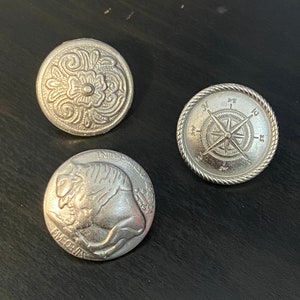 Puede incluir: Tres botones de metal plateados con diferentes dise&ntilde;os. El primer bot&oacute;n presenta un dise&ntilde;o floral, el segundo bot&oacute;n presenta un dise&ntilde;o de br&uacute;jula con las letras N, E, S y W, y el tercer bot&oacute;n presenta un dise&ntilde;o de b&uacute;falo con las palabras "IN GOD WE TRUST" y "FIVE CENTS" visibles.