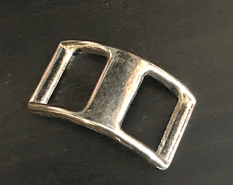 May include: A silver metal buckle with a curved shape and two rectangular openings.