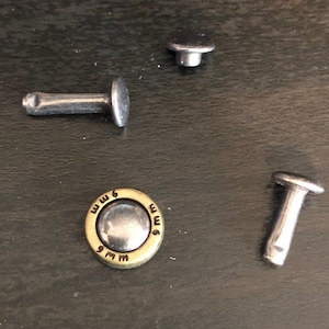 May include: Four silver metal rivets, one with a brass ring with the text "M 6 M" around the edge.
