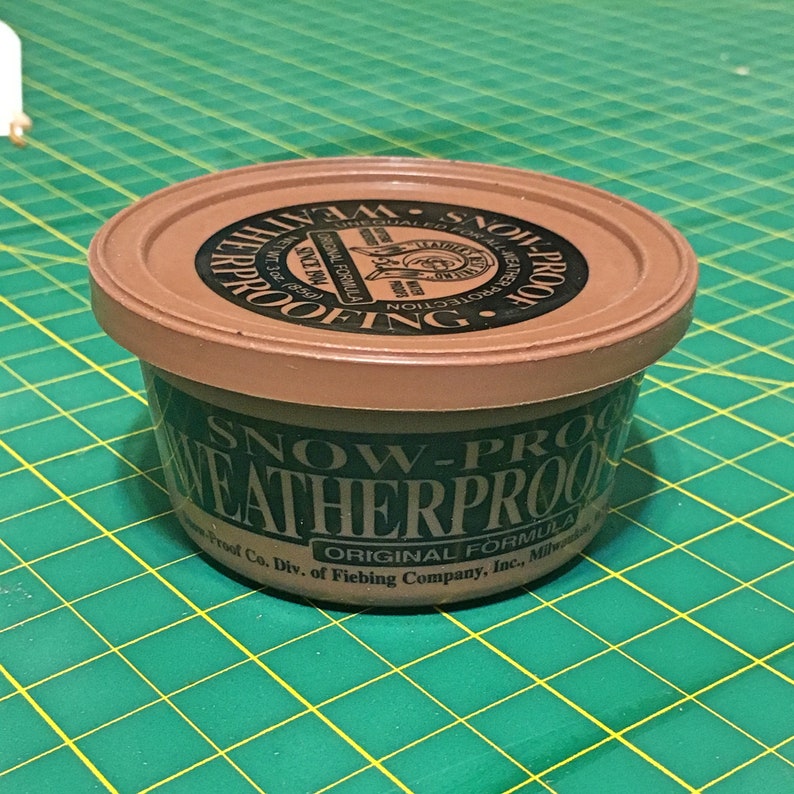 May include: A brown plastic container with a black lid. The lid has the text "Snow-Proof Leatherproofing" in a circle. The container has the text "Snow-Proof Weatherproof Original Formula" on the front. The container is sitting on a green surface.