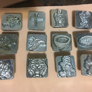 May include: Twelve silver metal stamps with various designs, including a cross, a cowboy hat, a bird, a rope, a paw print, and a bear. The stamps are arranged in a grid on a light brown surface.