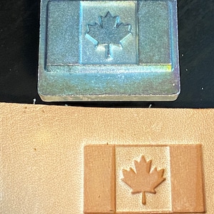 May include: A metal stamp with a design of the Canadian flag. The stamp is used to create an impression of the flag on leather.