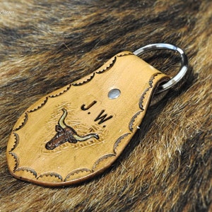 K&ouml;nnte beinhalten: Ein Schl&uuml;sselanh&auml;nger aus braunem Leder mit einem Stierkopfdesign und den eingepr&auml;gten Initialen "J.W.". Der Schl&uuml;sselanh&auml;nger hat einen silbernen Metallring.