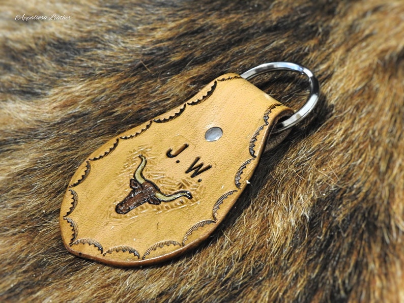 K&ouml;nnte beinhalten: Ein Schl&uuml;sselanh&auml;nger aus braunem Leder mit einem Stierkopfdesign und den eingepr&auml;gten Initialen "J.W.". Der Schl&uuml;sselanh&auml;nger hat einen silbernen Metallring.