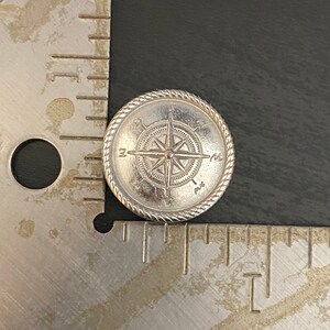 Puede incluir: Un amuleto de br&uacute;jula de plata con un dise&ntilde;o de cuerda alrededor del borde. La br&uacute;jula apunta al norte, sur, este y oeste.