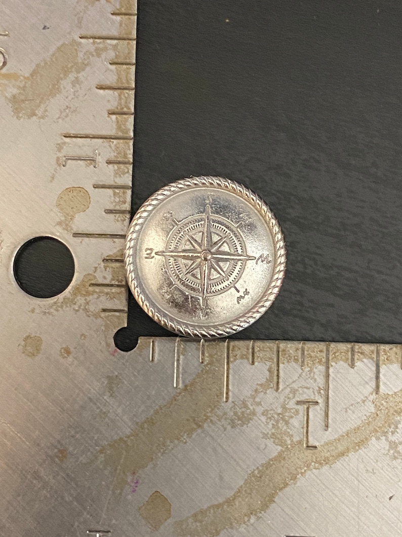 Puede incluir: Un amuleto de br&uacute;jula de plata con un dise&ntilde;o de cuerda alrededor del borde. La br&uacute;jula apunta al norte, sur, este y oeste.