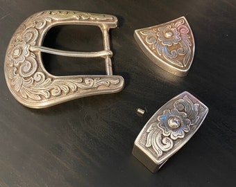 Puede incluir: Una hebilla de cinturón de plata con un diseño floral ornamentado. La hebilla está en tres piezas, con una hebilla principal, una hebilla más pequeña y un pasador. La hebilla está hecha de metal y tiene un acabado pulido.