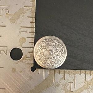 Puede incluir: Un bot&oacute;n de metal plateado con un dise&ntilde;o floral decorativo. El bot&oacute;n tiene un centro elevado y una superficie texturizada.