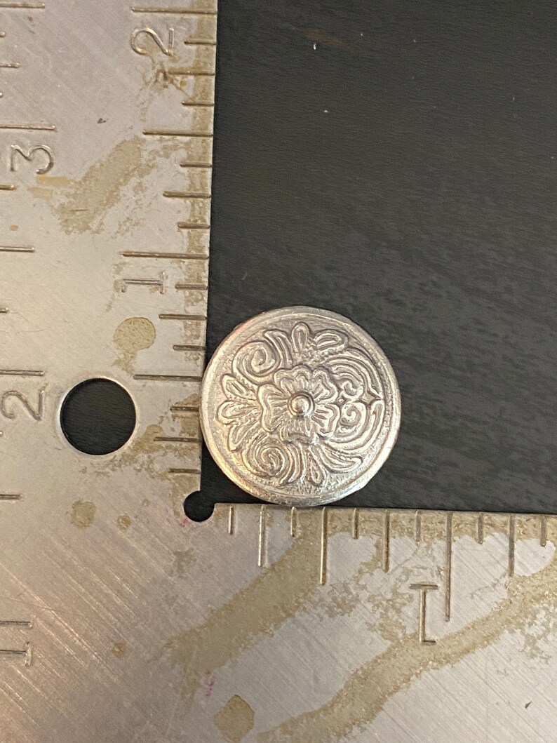 Puede incluir: Un bot&oacute;n de metal plateado con un dise&ntilde;o floral decorativo. El bot&oacute;n tiene un centro elevado y una superficie texturizada.