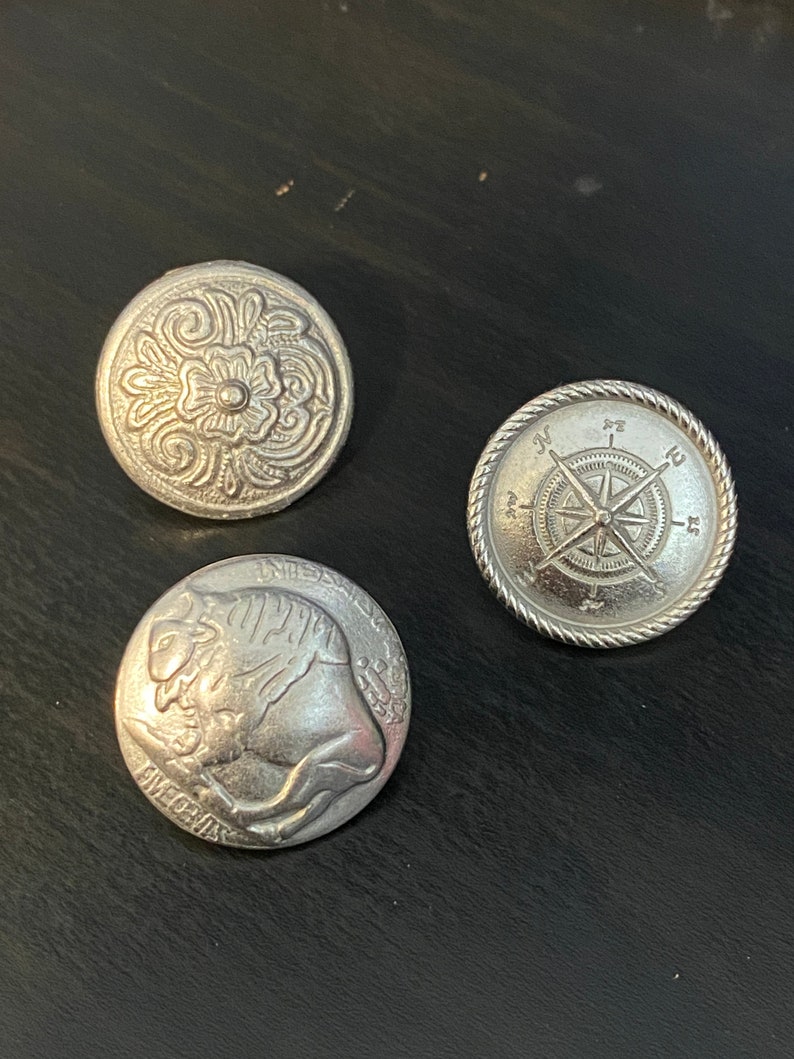 Puede incluir: Tres botones de metal plateados con diferentes dise&ntilde;os. El primer bot&oacute;n presenta un dise&ntilde;o floral, el segundo bot&oacute;n presenta un dise&ntilde;o de br&uacute;jula con las letras N, E, S y W, y el tercer bot&oacute;n presenta un dise&ntilde;o de b&uacute;falo con las palabras "IN GOD WE TRUST" y "FIVE CENTS" visibles.