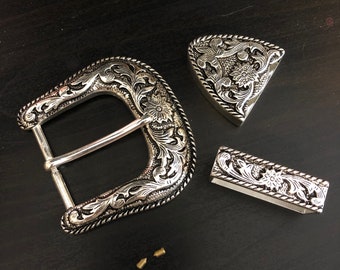 May include: A silver belt buckle set with a floral design. The set includes a buckle, a tip, and a keeper. The buckle is a traditional western style with a large, ornate design. The tip and keeper are smaller and more simple, but still feature the same floral design.