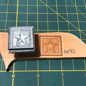 May include: A silver metal stamp with a five-pointed star design inside a square frame. The stamp is used to create an impression on a piece of brown leather. The leather has the number 8672 written on it.