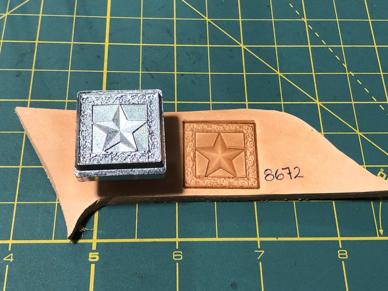 May include: A silver metal stamp with a five-pointed star design inside a square frame. The stamp is used to create an impression on a piece of brown leather. The leather has the number 8672 written on it.