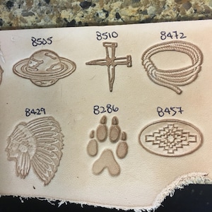 May include: A close-up of a leather surface with various leather stamping designs. The designs include a planet, a nail, a rope, a headdress, a paw print, and a geometric pattern. Each design is labeled with a number.