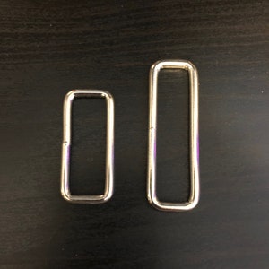 May include: Two rectangular metal rings with rounded corners. The rings are silver in color and appear to be made of a shiny metal. One ring is smaller than the other. The rings are placed on a dark surface.
