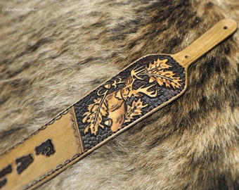 May include: A brown leather rifle sling with a tooled design of a deer in the woods. The sling has a brown leather strap with a hole at the end for attaching to a rifle.