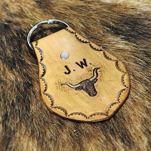 K&ouml;nnte beinhalten: Ein brauner Leder-Schl&uuml;sselanh&auml;nger mit einem dekorativen, gewellten Rand. Der Schl&uuml;sselanh&auml;nger zeigt ein gepr&auml;gtes Design eines Longhorn-Stiers mit den Initialen "J.W." dar&uuml;ber.