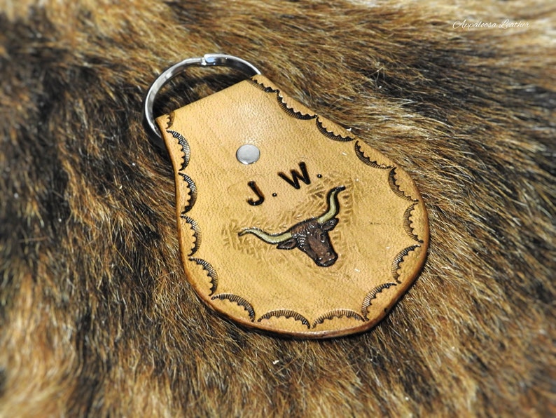 K&ouml;nnte beinhalten: Ein brauner Leder-Schl&uuml;sselanh&auml;nger mit einem dekorativen, gewellten Rand. Der Schl&uuml;sselanh&auml;nger zeigt ein gepr&auml;gtes Design eines Longhorn-Stiers mit den Initialen "J.W." dar&uuml;ber.