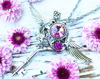 May include: A silver key pendant necklace with a pink crystal and ornate wings. The necklace is surrounded by pink flowers.