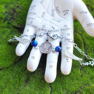 May include: A silver chain bracelet featuring a vintage coin, blue beads, and wing charms. The bracelet is displayed on a white hand sculpture against a green, mossy background. The bracelet's chain is approximately 7 inches long.