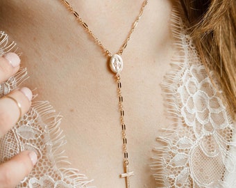 Puede incluir: Un collar de cadena dorada con un pequeño colgante ovalado y un dije de cruz. El collar lo lleva una persona con el pelo largo y castaño y una blusa de encaje blanca.