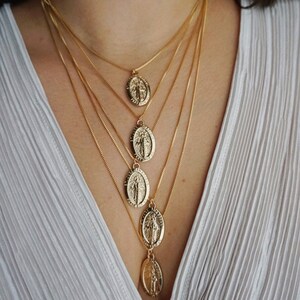 K&ouml;nnte beinhalten: Eine mehrreihige goldene Kettenkette mit f&uuml;nf ovalen Anh&auml;ngern, die ein religi&ouml;ses Symbol zeigen. Die Kette wird von einer Person mit einem wei&szlig;en Hemd getragen.