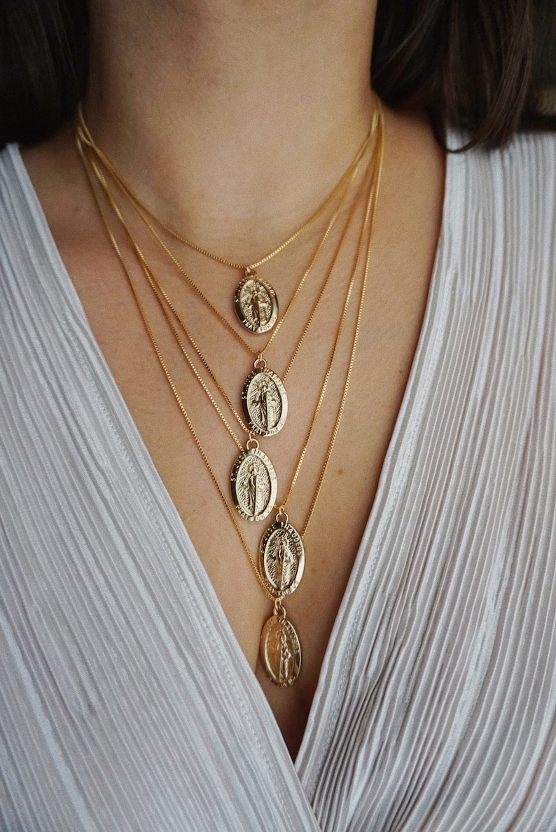 K&ouml;nnte beinhalten: Eine mehrreihige goldene Kettenkette mit f&uuml;nf ovalen Anh&auml;ngern, die ein religi&ouml;ses Symbol zeigen. Die Kette wird von einer Person mit einem wei&szlig;en Hemd getragen.