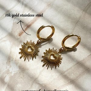 May include: Gold hoop earrings with a sunburst charm featuring a heart cutout. The earrings are made of 18k gold stainless steel and are waterproof and hypoallergenic.