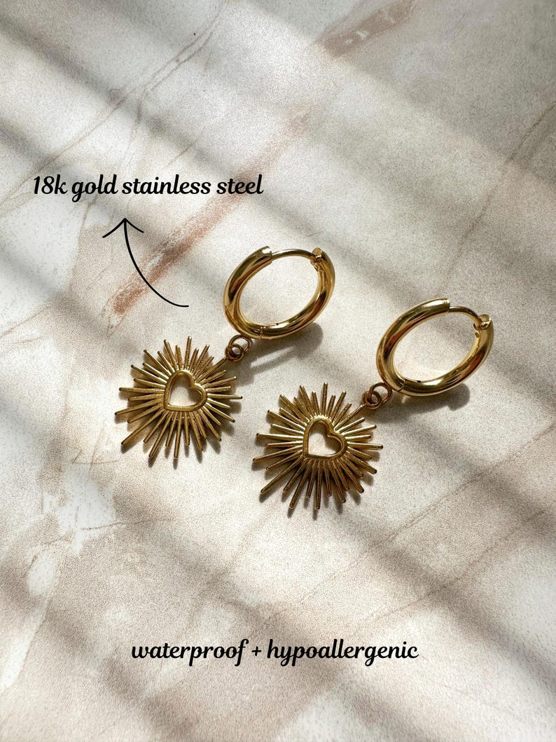 May include: Gold hoop earrings with a sunburst charm featuring a heart cutout. The earrings are made of 18k gold stainless steel and are waterproof and hypoallergenic.