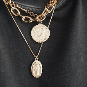 K&ouml;nnte beinhalten: Drei goldene Halsketten mit Anh&auml;ngern. Die oberste Halskette ist eine dicke Kette mit einem gro&szlig;en runden M&uuml;nzanh&auml;nger. Die mittlere Halskette ist eine d&uuml;nnere Kette mit einem kleineren runden M&uuml;nzanh&auml;nger. Die unterste Halskette ist eine zarte Kette mit einem ovalen Anh&auml;nger.