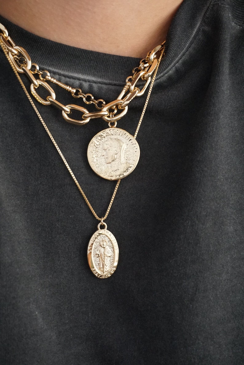 K&ouml;nnte beinhalten: Drei goldene Halsketten mit Anh&auml;ngern. Die oberste Halskette ist eine dicke Kette mit einem gro&szlig;en runden M&uuml;nzanh&auml;nger. Die mittlere Halskette ist eine d&uuml;nnere Kette mit einem kleineren runden M&uuml;nzanh&auml;nger. Die unterste Halskette ist eine zarte Kette mit einem ovalen Anh&auml;nger.