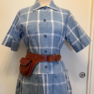 May include: A vintage blue and white plaid dress with short sleeves and a button-down front. A brown leather belt with a small pouch is worn at the waist. The dress is displayed on a mannequin.