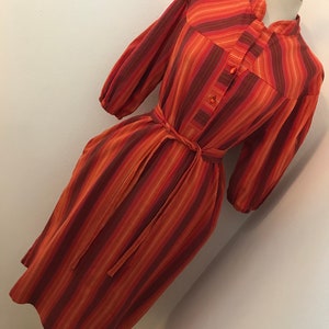 May include: A long-sleeved, orange and brown striped dress with a button-up front and a tie belt. The dress has a mandarin collar and a slightly puffed sleeve.