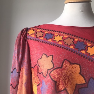May include: Red, orange, yellow, and blue floral print blouse with a boat neck and long sleeves.