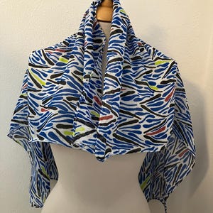 May include: A blue and white scarf with a bold abstract pattern of blue, black, red, and yellow brushstrokes. The scarf is draped over a mannequin, showcasing its flowing design. This accessory adds a pop of color.