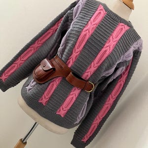 May include: A gray cable-knit sweater with pink and lavender accents. The sweater features vertical pink cable-knit stripes and lavender cable-knit sections on the sleeves. A brown leather belt with a gold buckle and a small brown leather pouch are worn around the waist.