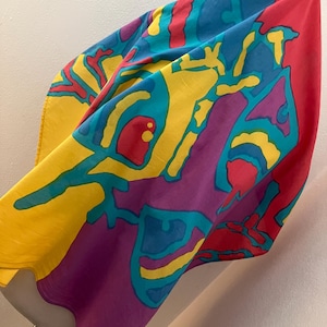 May include: A colorful silk scarf with an abstract design. The scarf features a vibrant mix of colors, including yellow, blue, red, and purple. The design has a unique, artistic pattern. The scarf is draped over a mannequin, showcasing its shape and texture.