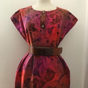 Puede incluir: Un vestido de estampado floral rosa y naranja con un cinturón marrón. El vestido tiene un escote redondo y mangas cortas.